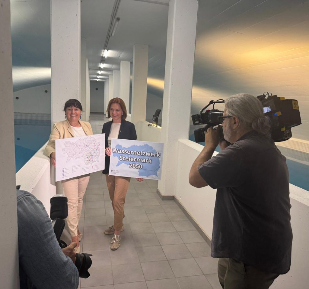 Zwei Frauen stehen in einer modernen Wasseraufbereitungsanlage und halten gemeinsam Schilder in die Kamera – eines mit einer Landkarte und das andere mit der Aufschrift ‚Wassernetzwerk Steiermark 2050‘. Im Hintergrund ein Wasserbecken, im Vordergrund ein Kamerateam.